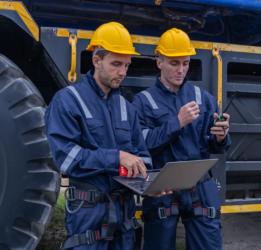 Two workers in hard hats using a laptop beside heavy machinery