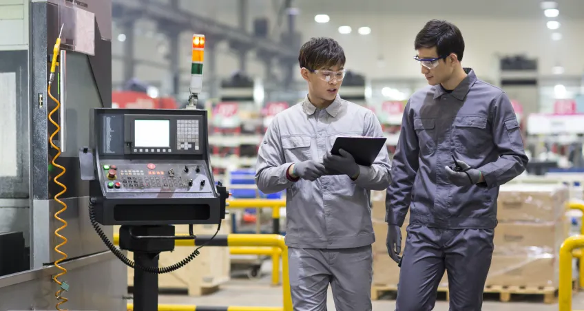 Two workers at a machine control panel with a tablet