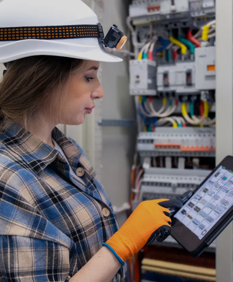Engineer in a hard hat inspecting an electrical control panel with a tablet