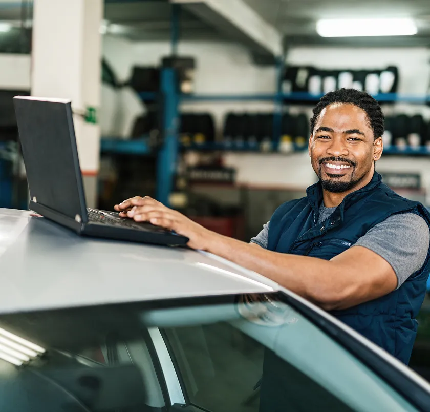 Smiling mechanic using a laptop resting on a car in a workshop