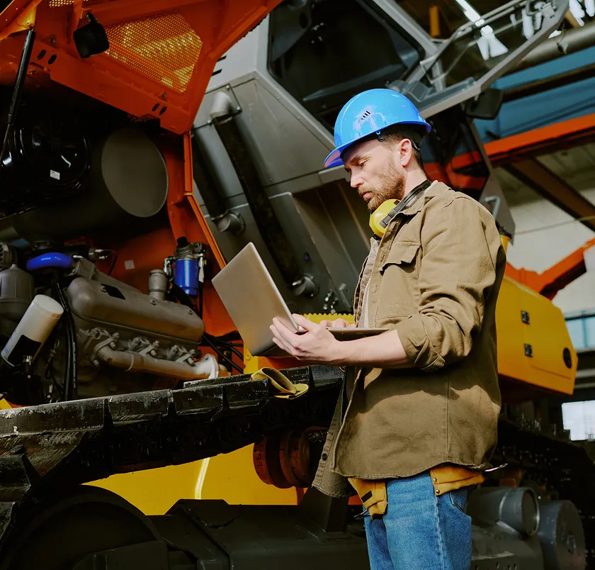 Engineer in a hard hat using a laptop on yellow plant machinery
