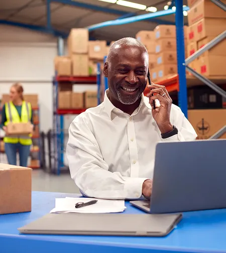 Smiling man on the phone using a laptop in a warehouse