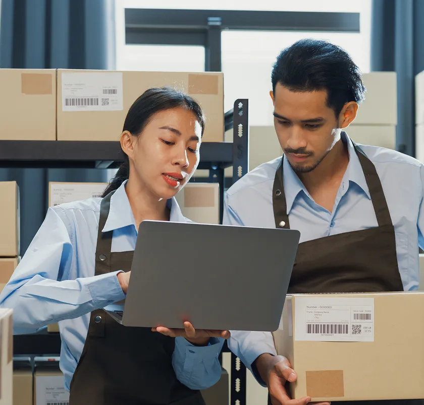Two workers in aprons reviewing orders on a laptop beside labelled parcels