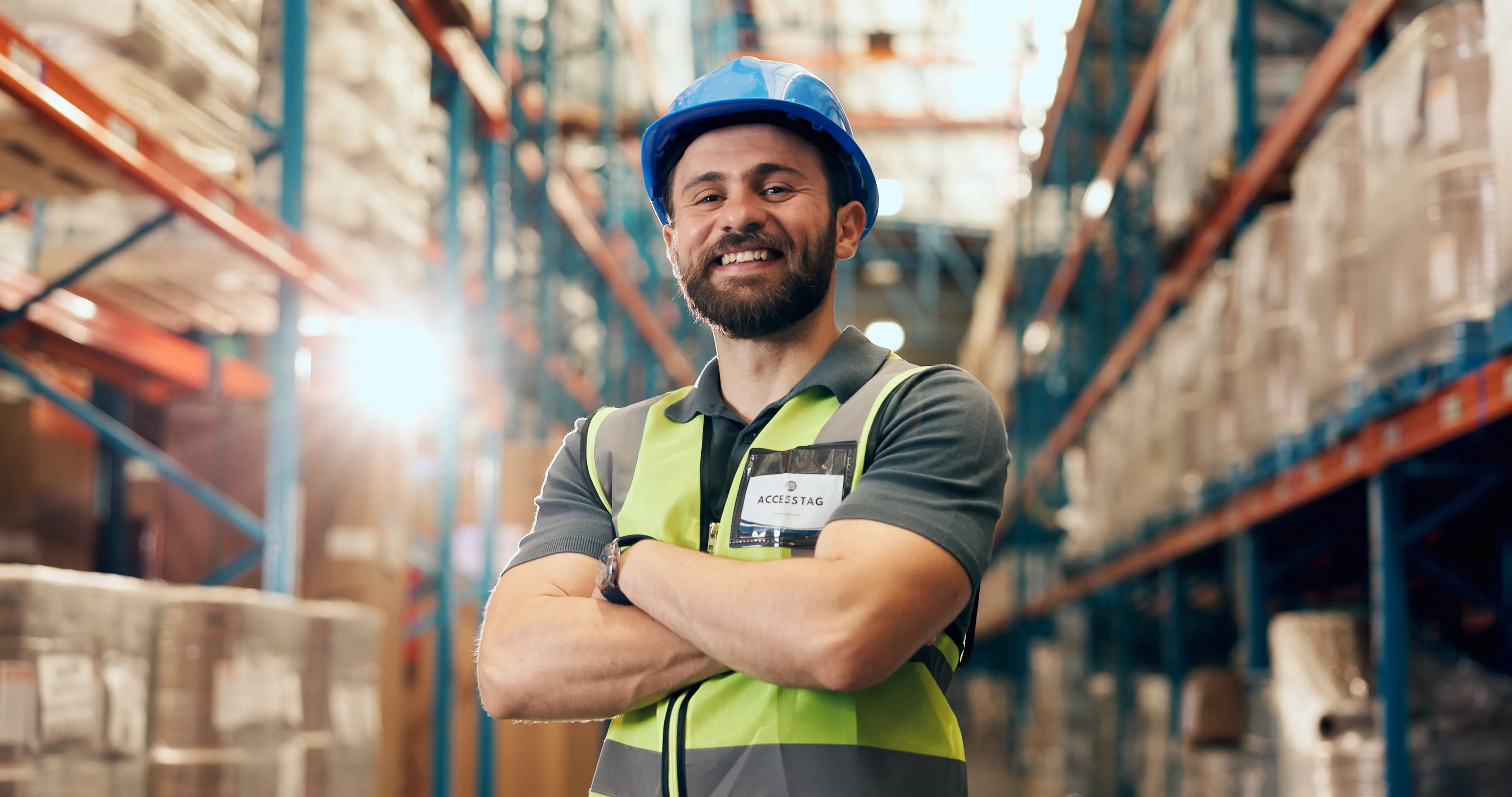 Smiling warehouse worker wearing a blue hard hat and neon safety vest with arms crossed.