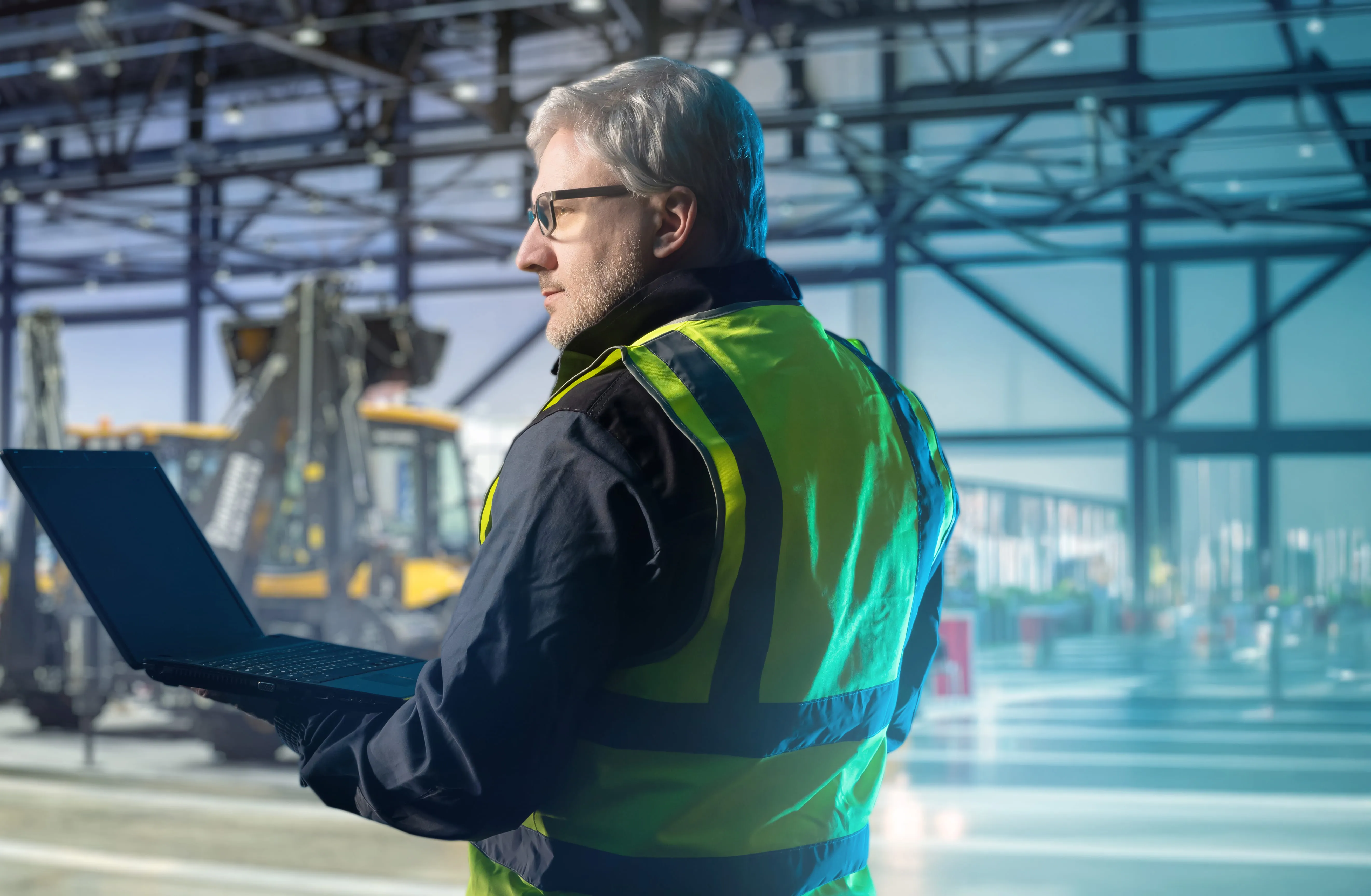 Man in safety vest using laptop in industrial warehouse with heavy machinery in background.