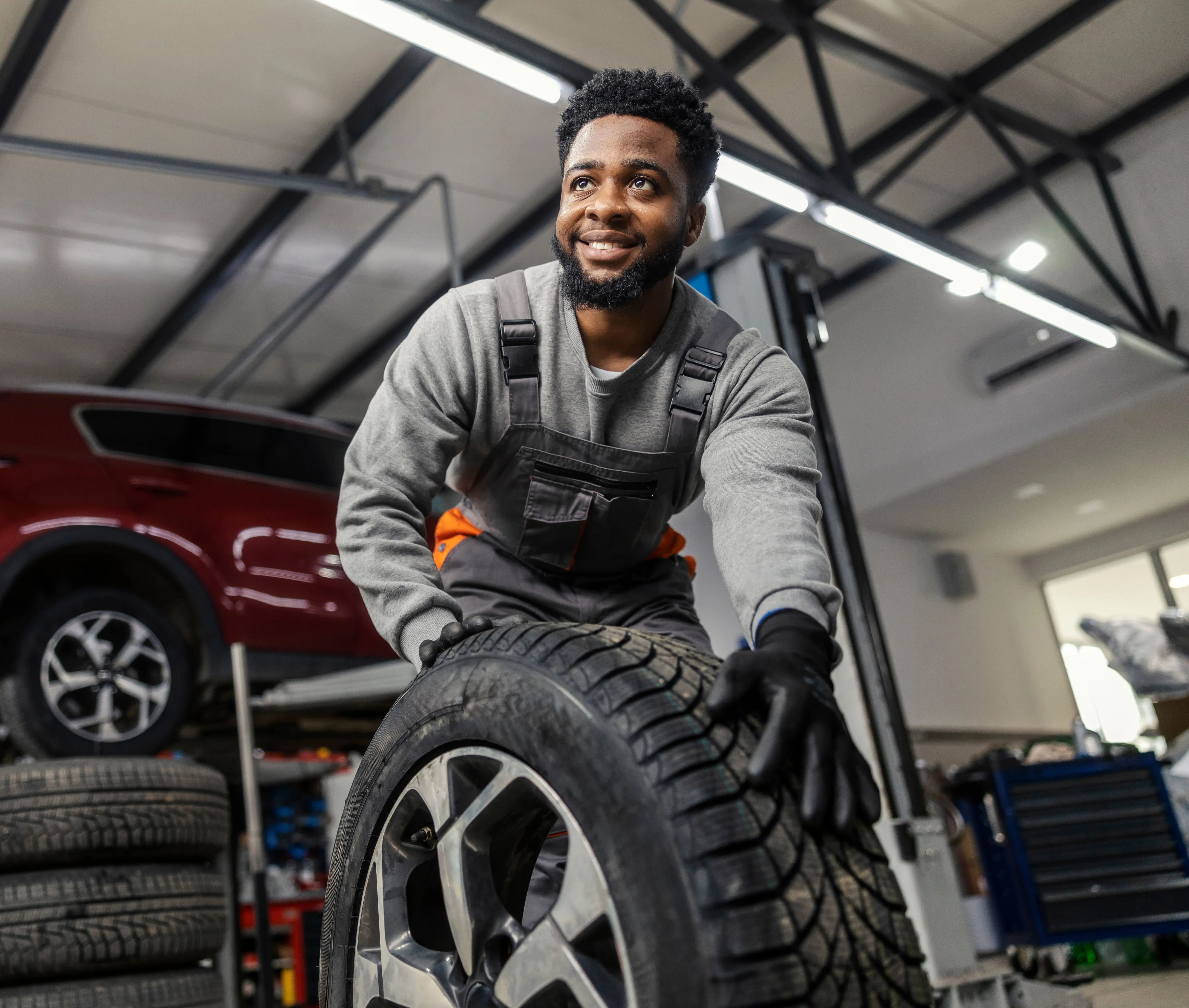 Smiling mechanic in gray overalls holding a car tire in an auto repair shop with a red car nearby.