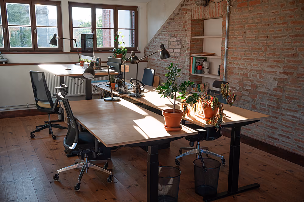 Modernes Büro mit Holztischen, Schreibtischlampen, Bürostühlen und Zimmerpflanzen an der Wand mit Ziegelsteinen.