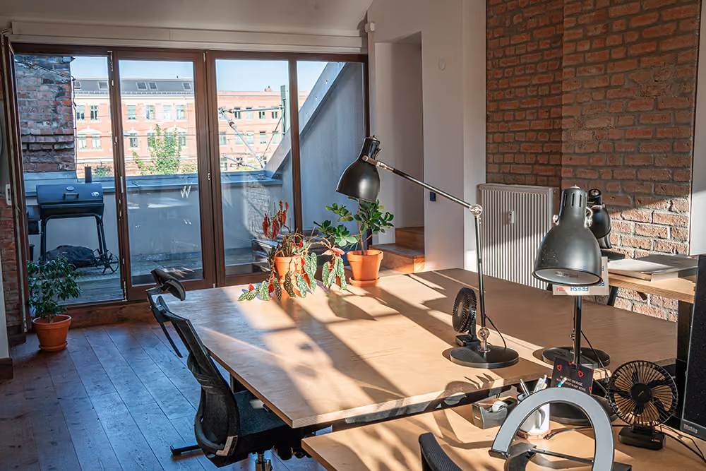 Sonniger Arbeitsplatz mit Holzschreibtisch, Pflanzen und schwarzen Lampen neben Fenster zur Terrasse.