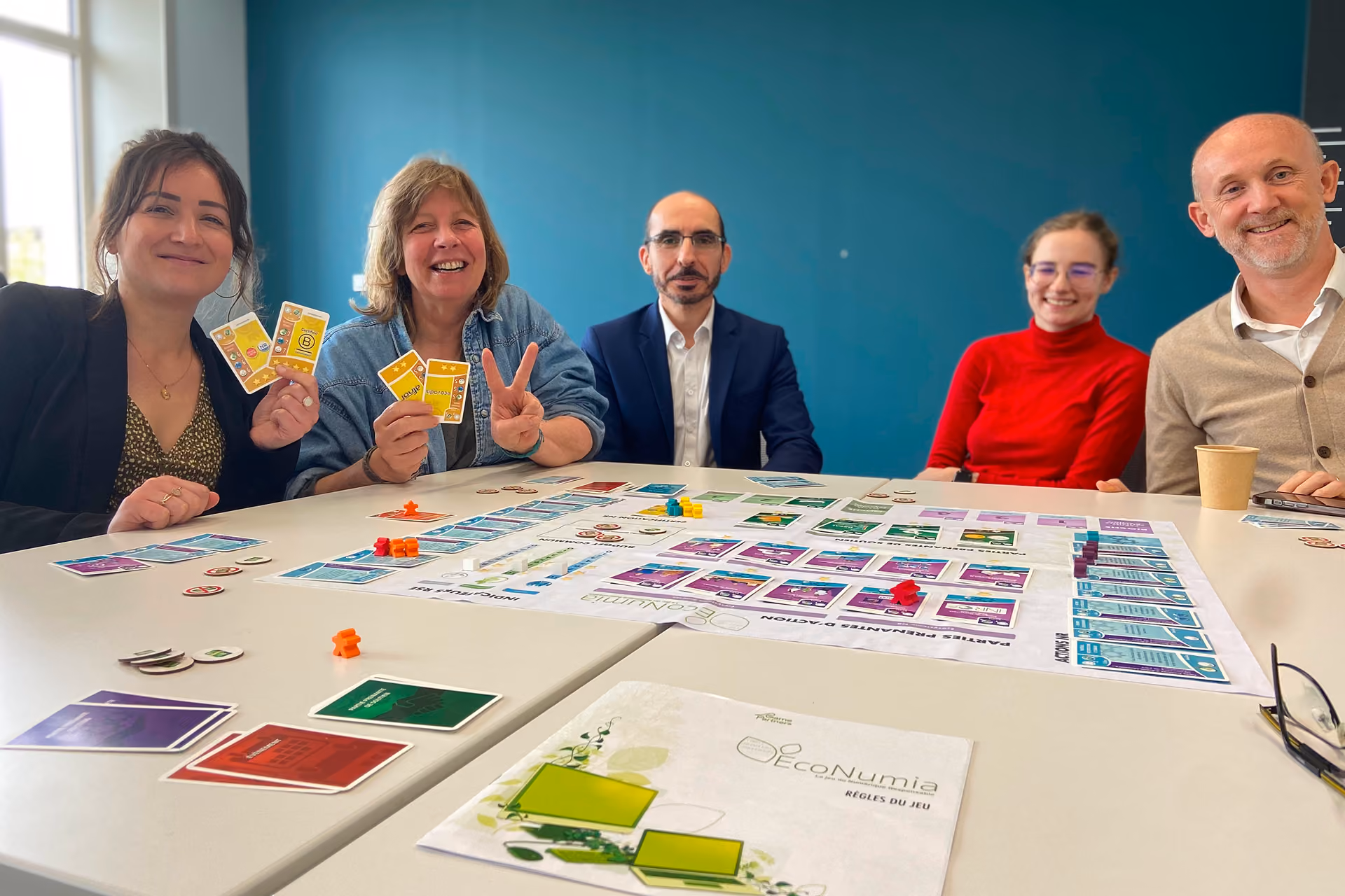 Groupe de participants souriants en fin de partie d'ECONUMIA, plateau et règles du jeu sur la table