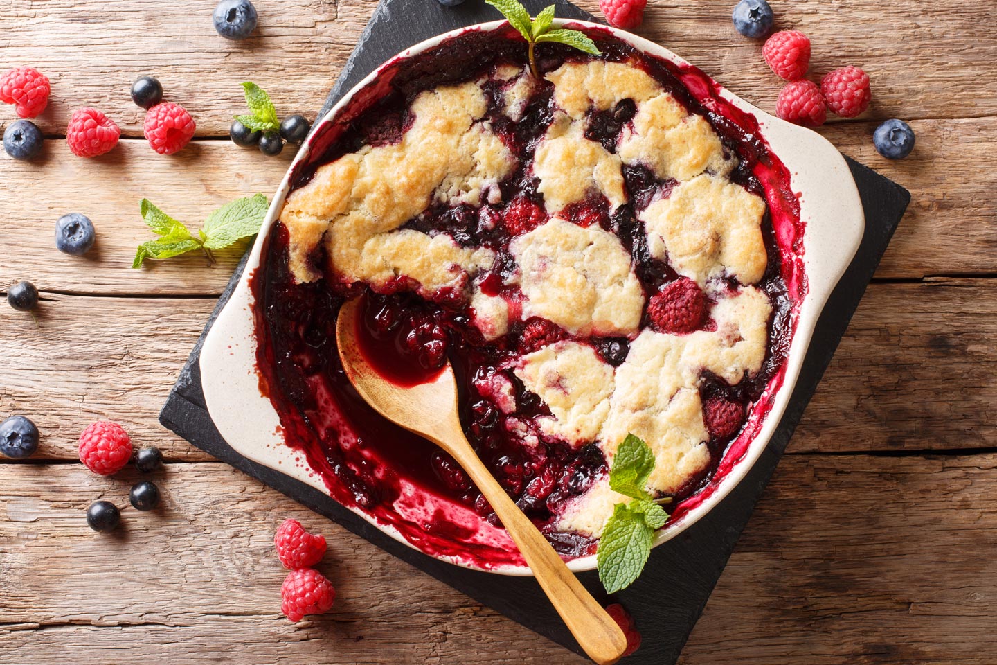 Mixed berry cobbler in a baking dish with a wooden spoon and garnished with mint leaves on rustic wood.