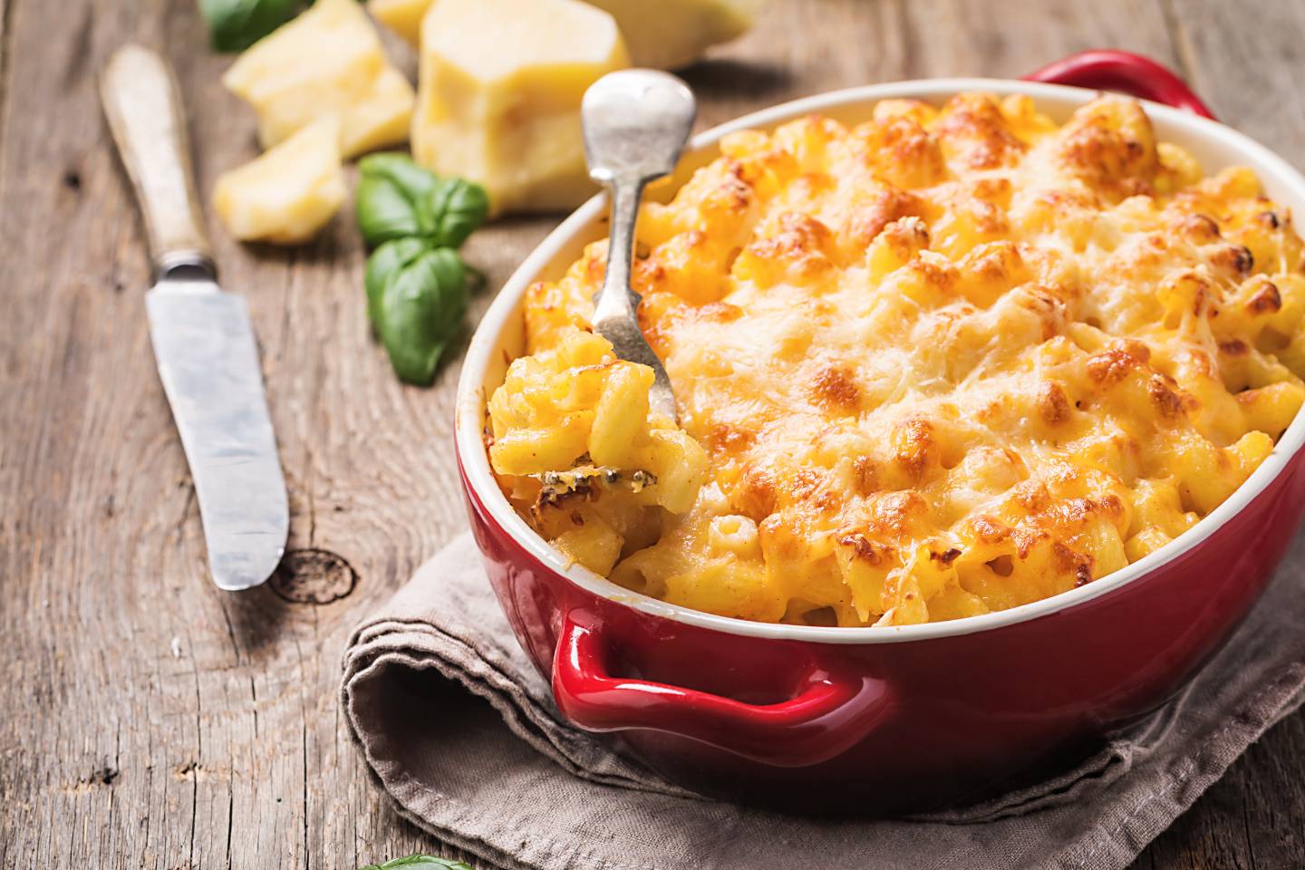 Baked macaroni and cheese in a red dish on a cloth with a knife and cheese chunks nearby.