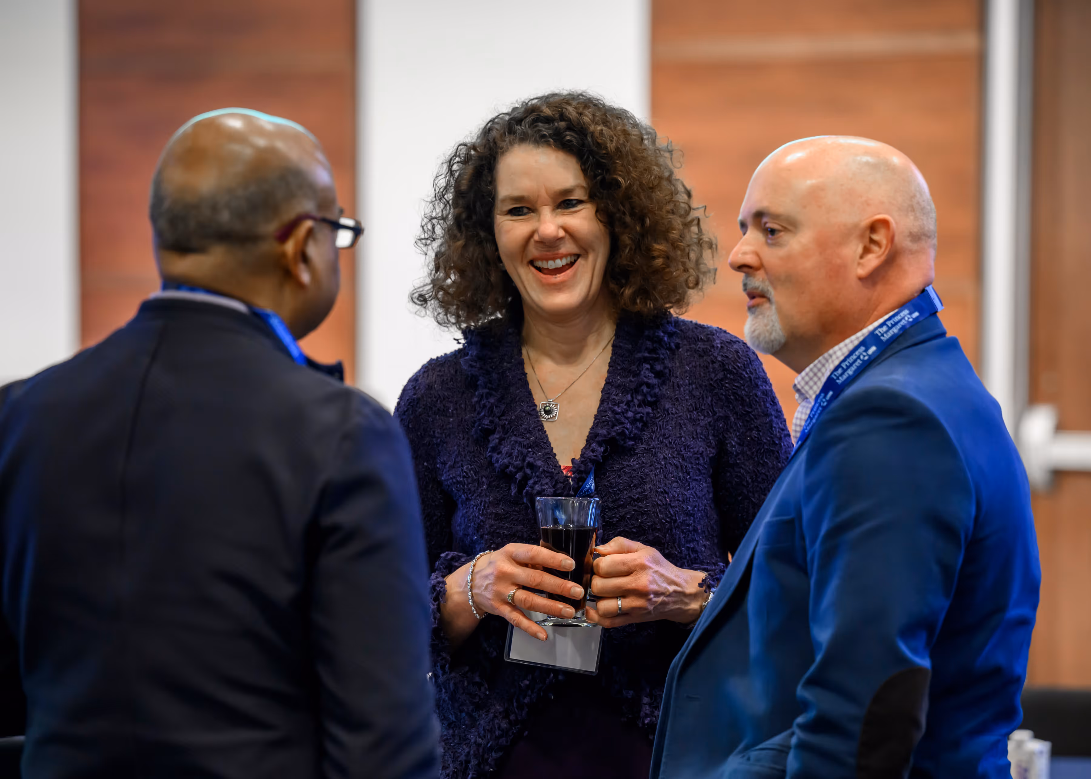 Three professionals in conversation, woman in purple holding a drink, two men in suits with lanyards.
