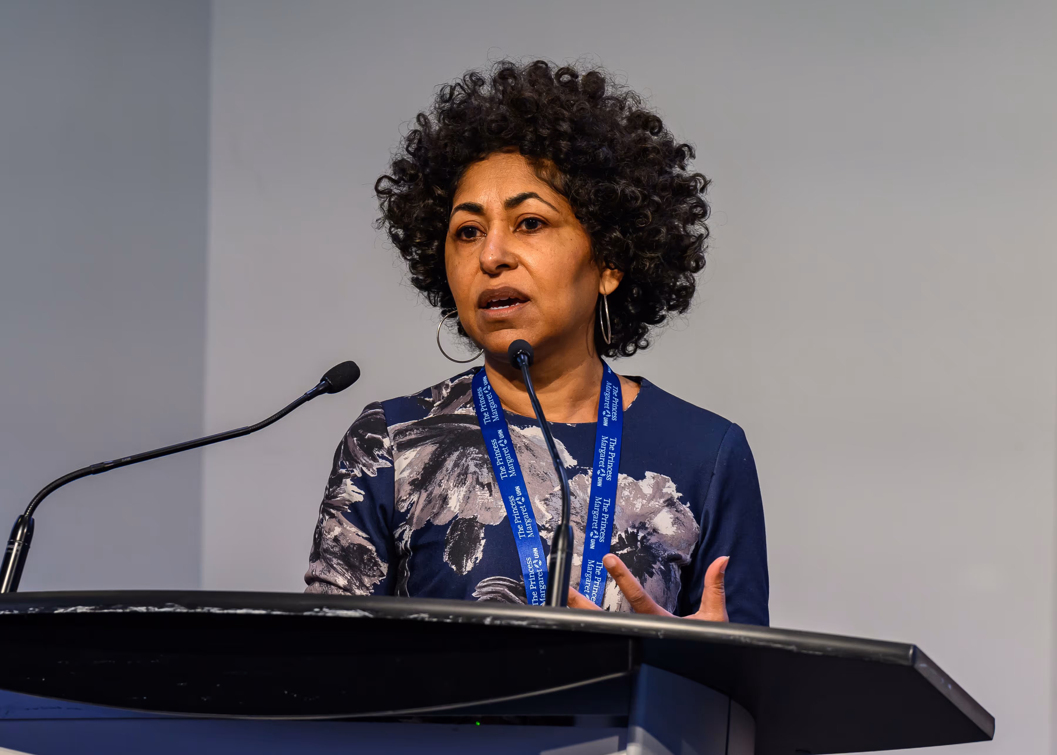 Woman with curly hair speaking at a podium with microphones and a conference lanyard.