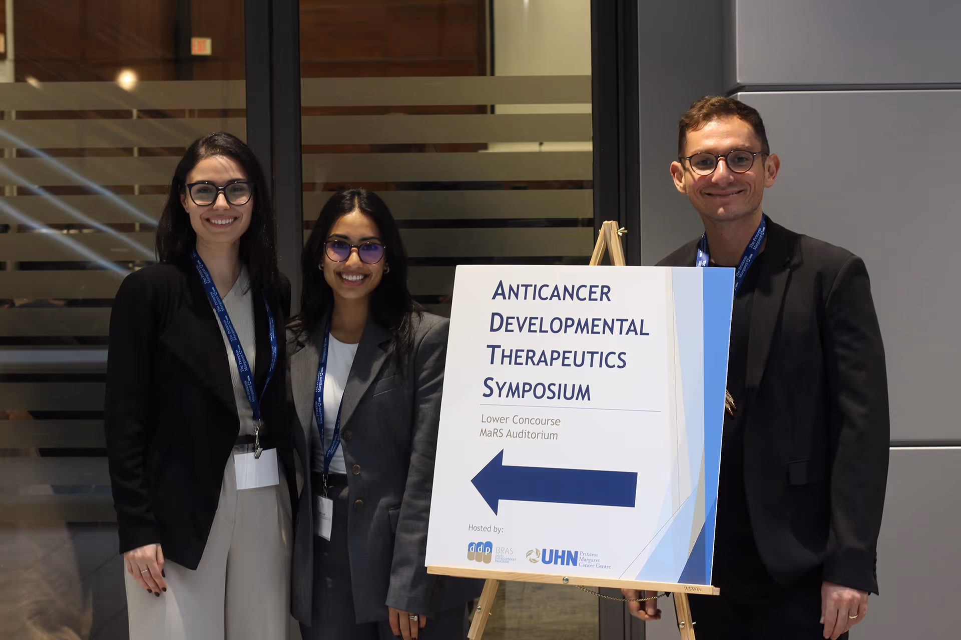 Three people with conference badges stand next to a sign for the Anticancer Developmental Therapeutics Symposium.