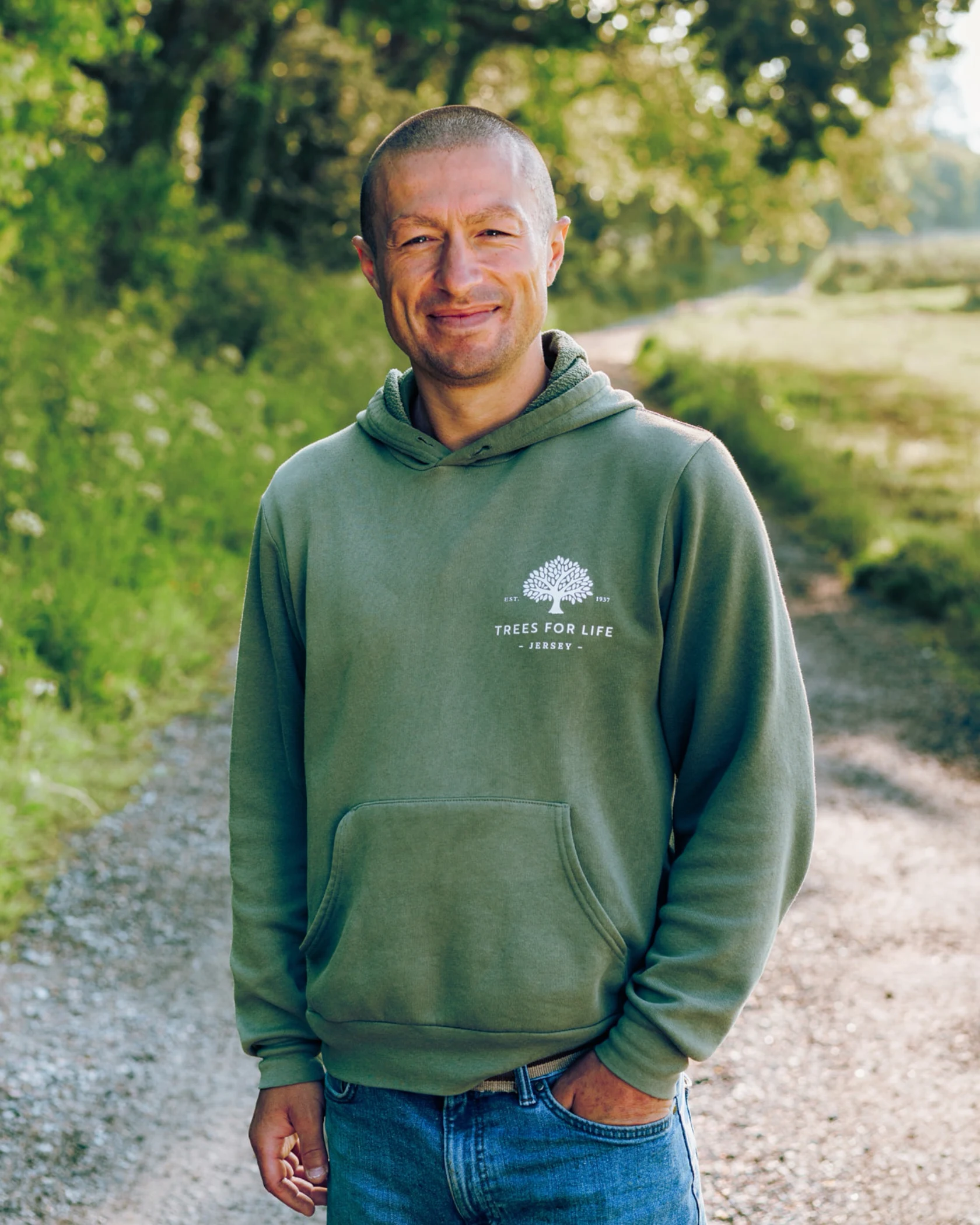 Smiling man in green hoodie and jeans standing on a forest path with greenery in background.