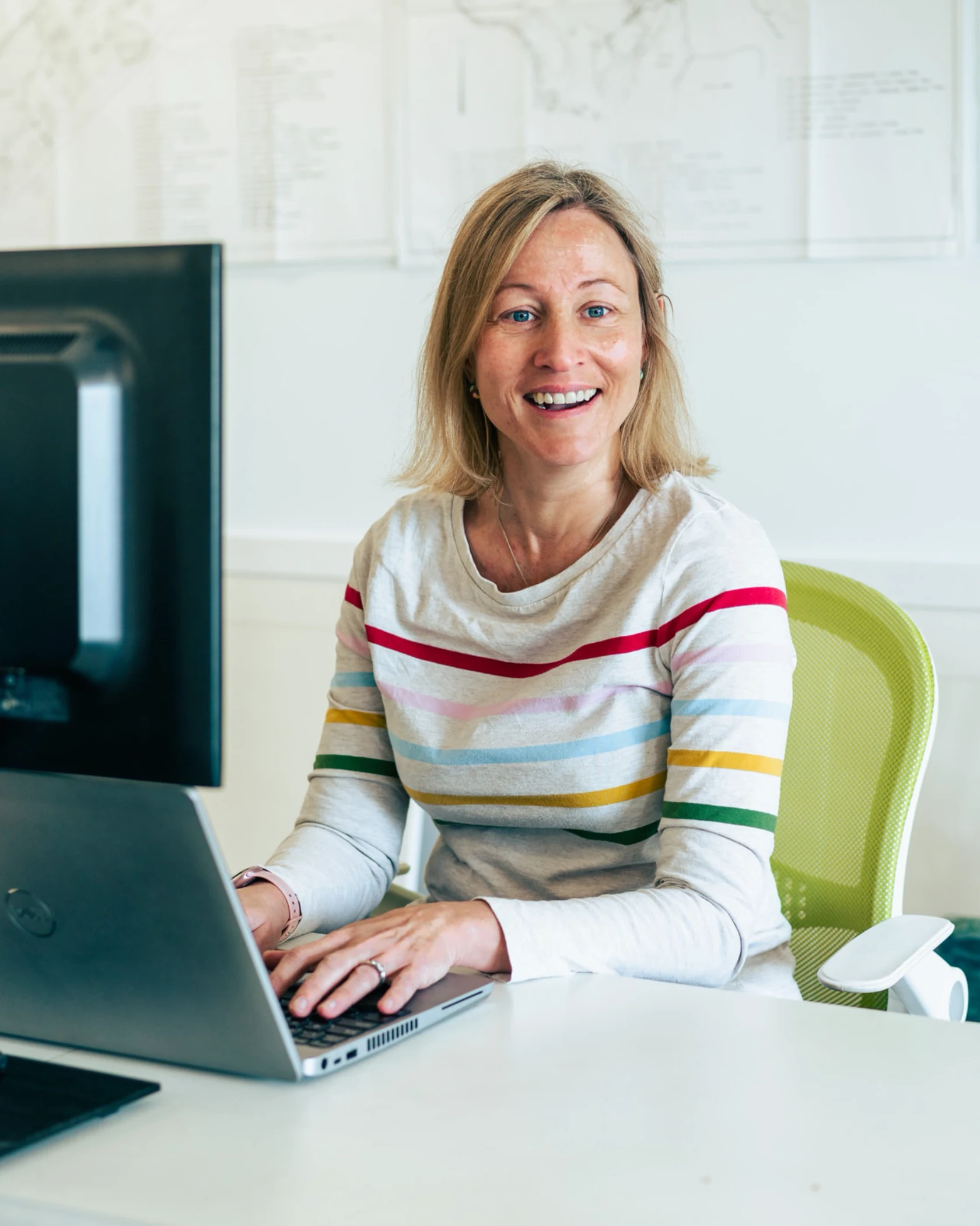 Smiling woman wearing a striped shirt sitting at a desk working on a laptop.
