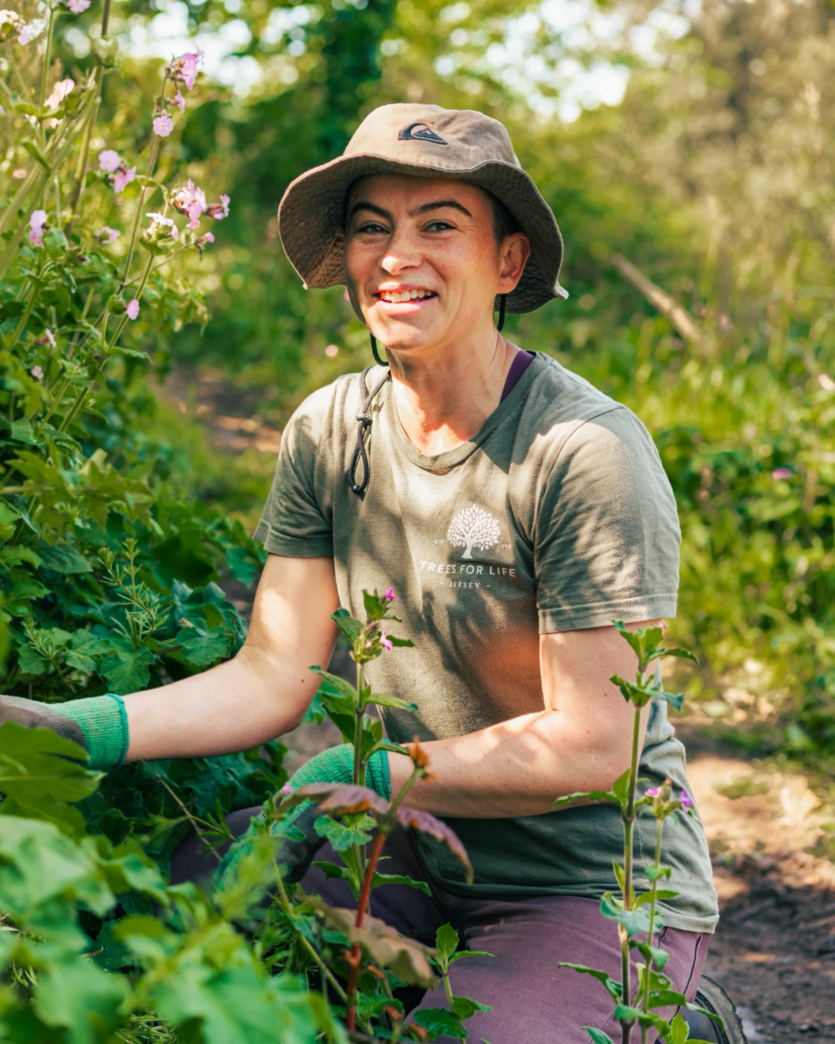 Person wearing a bucket hat and green gloves tending plants in a garden on a sunny day.