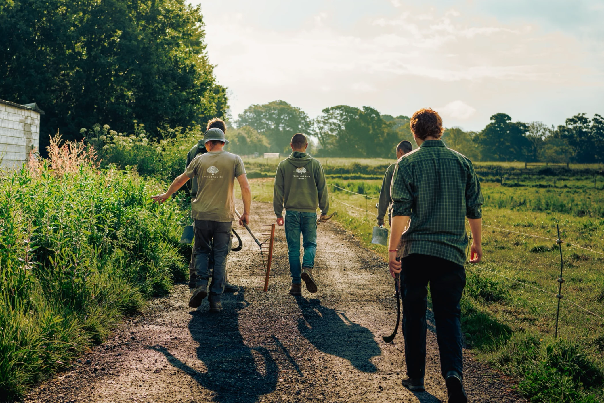 Group of five people walking on a rural path in green fields carrying gardening tools.