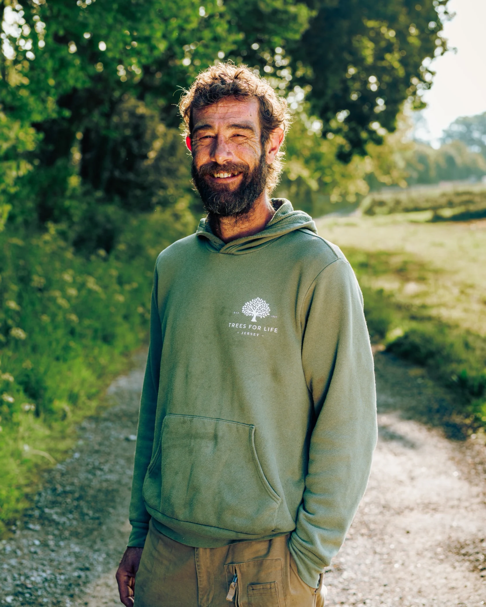 Smiling man with beard wearing a green 'Trees for Life Jersey' hoodie outdoors on a sunny pathway.