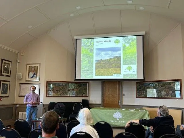 Presenter at a lecture showing a slide titled 'Egypte Woods' to an audience in a room.
