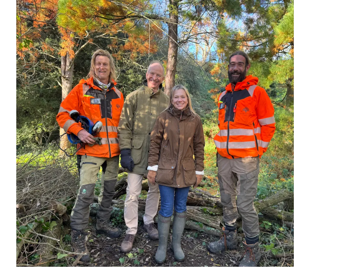 Four people standing outdoors in fall foliage, two wearing bright orange safety jackets and work boots.