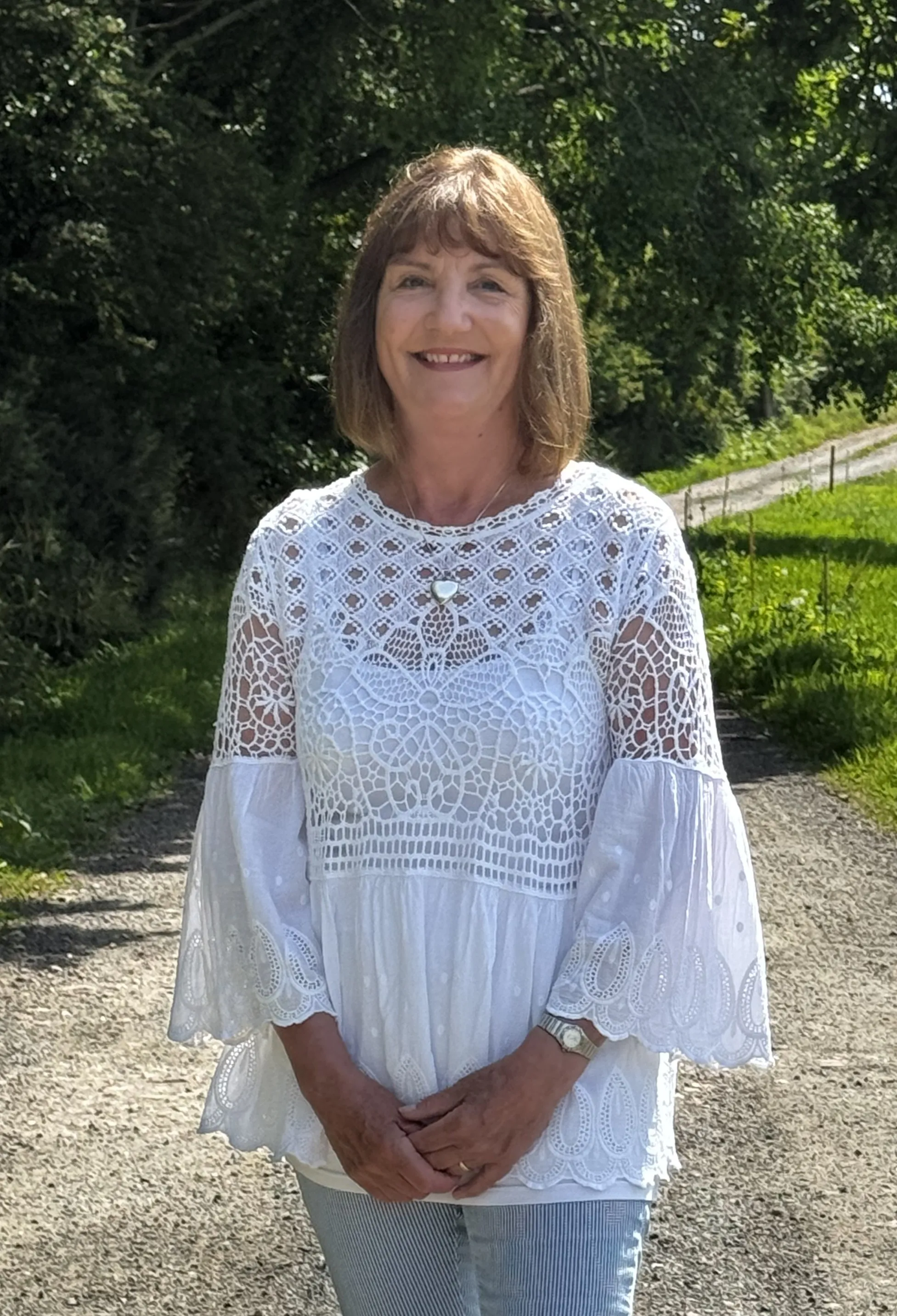 Smiling woman in a white lace top and striped pants stands on a sunlit gravel path with greenery.