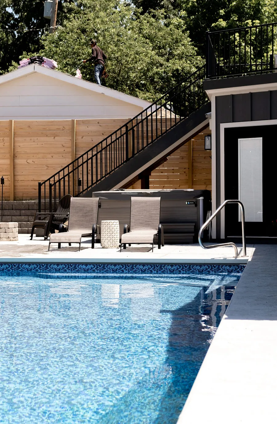 Man working on a roof behind poolside lounge chairs beside a hot tub and stairs.