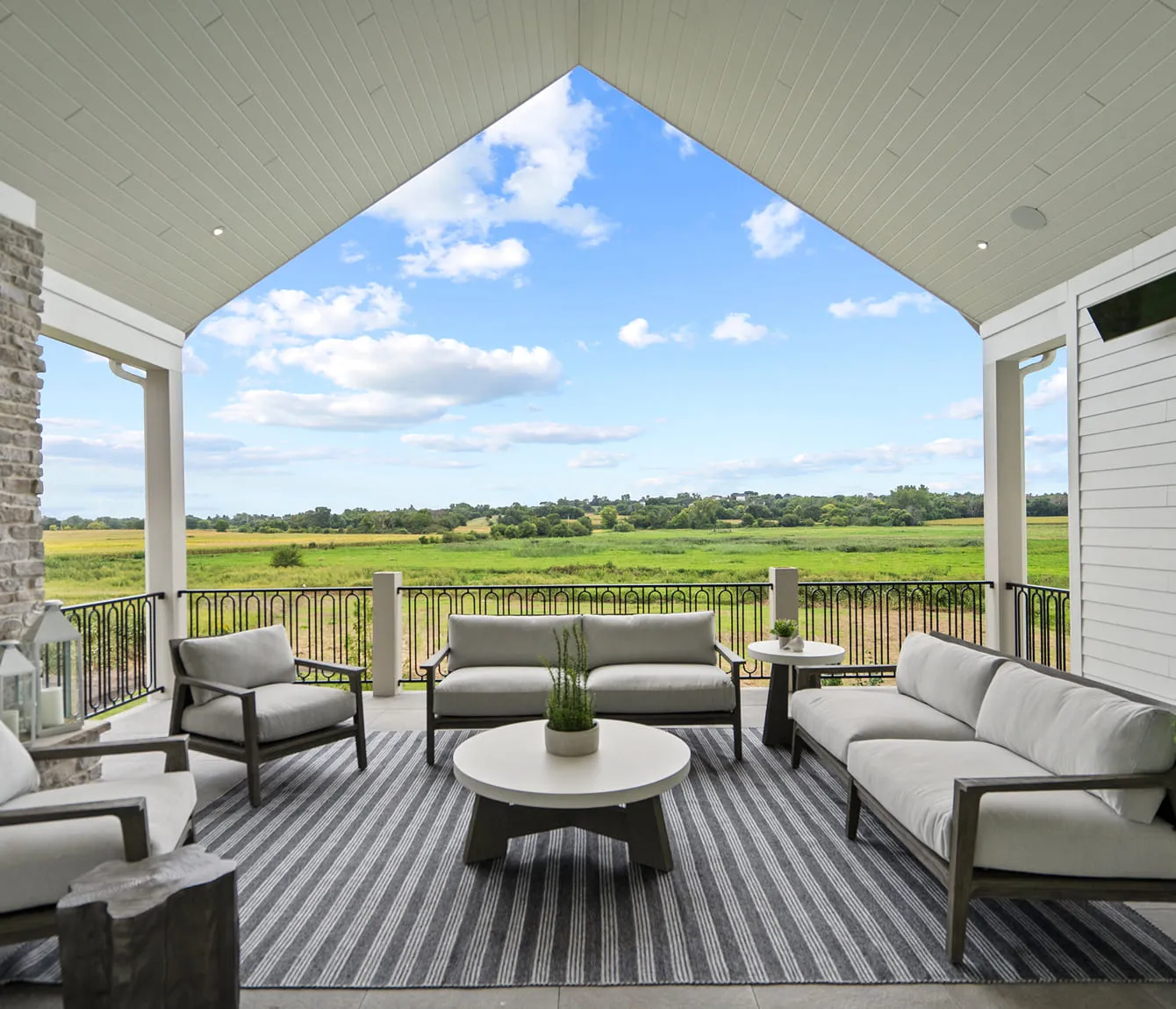 Covered patio with modern outdoor sofas, coffee table, and green field view under a blue sky.