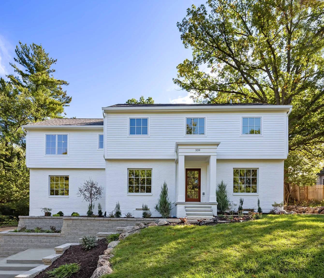Two-story white house with large windows, front porch, steps, and landscaped yard with trees and grass.
