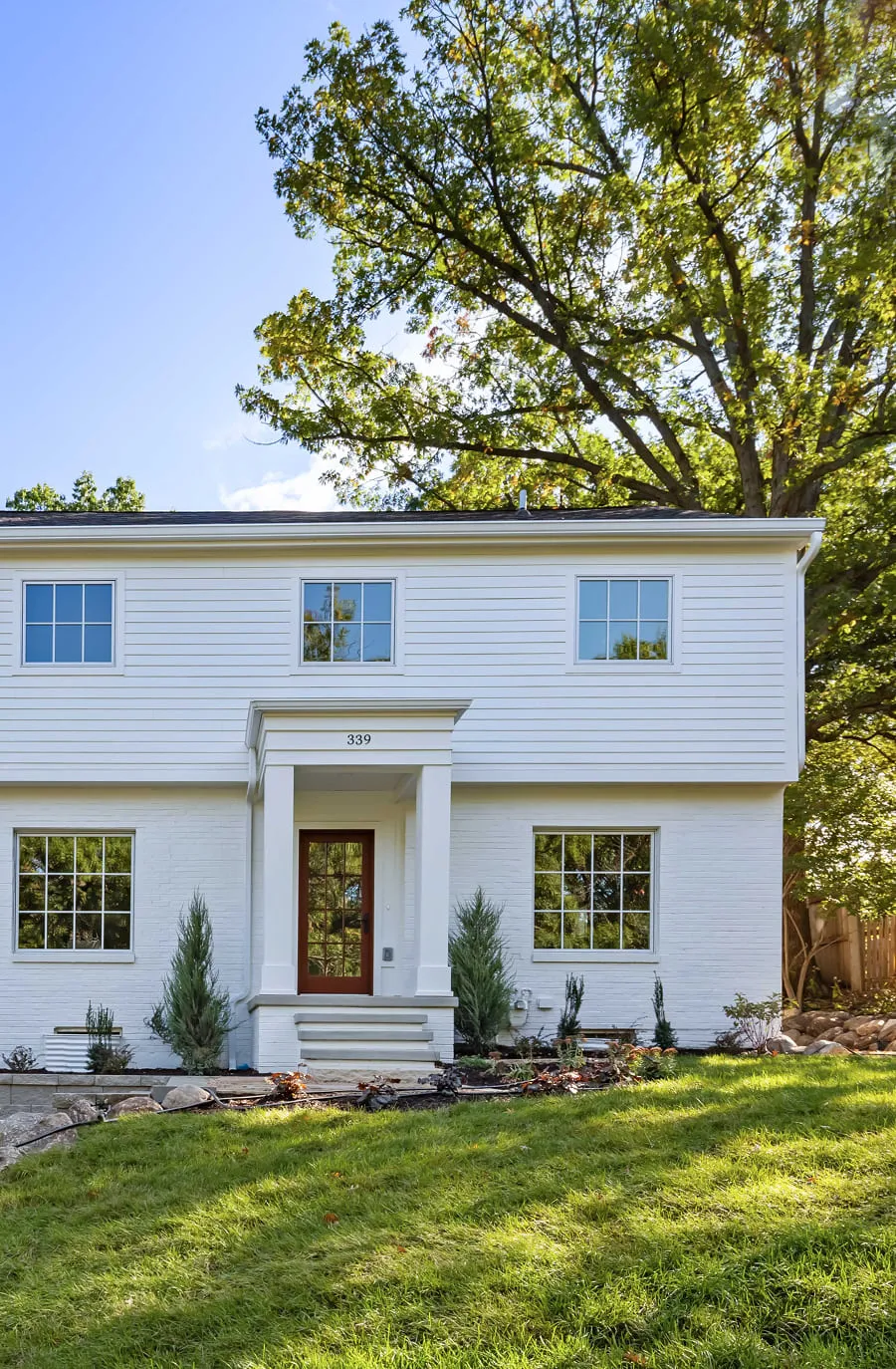 White two-story house with a wooden door, large windows, and green lawn in front under a tree.