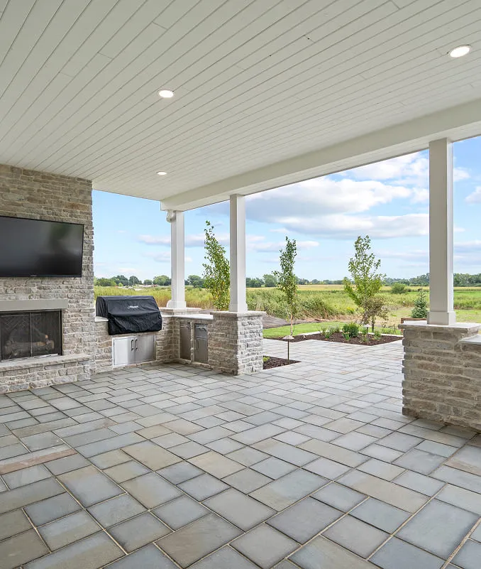 Covered outdoor patio with stone fireplace, built-in grill, TV, and view of landscaped yard and fields.