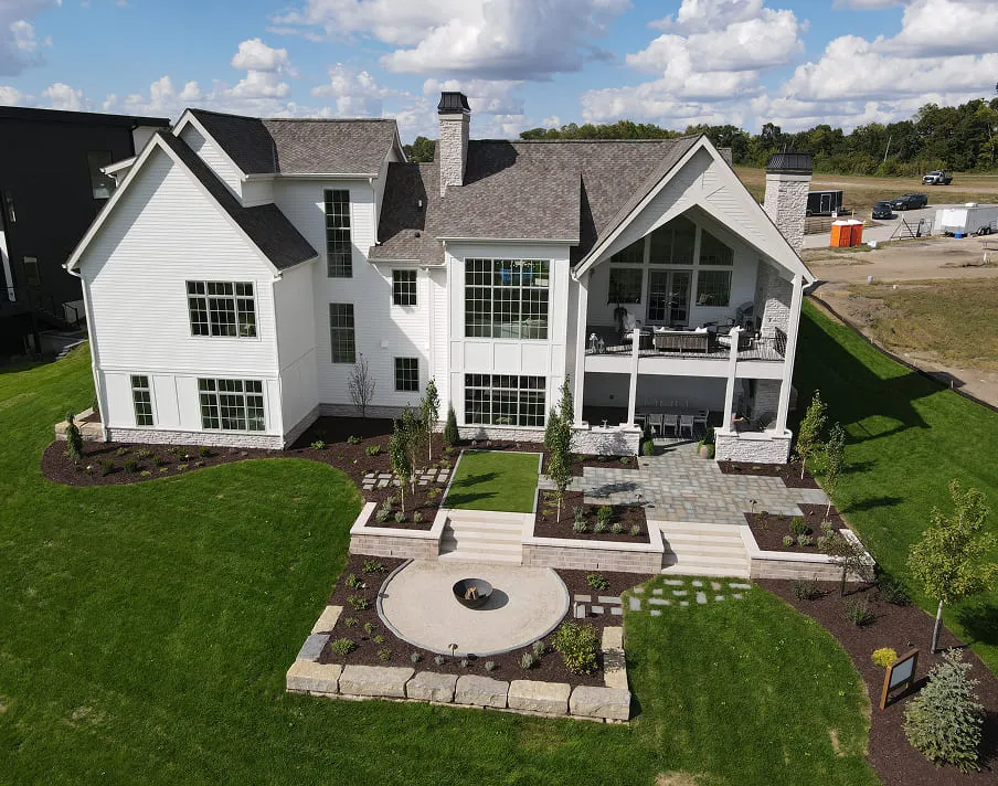 Large white house with multiple windows, a patio, landscaped yard, and fire pit in a grassy area.