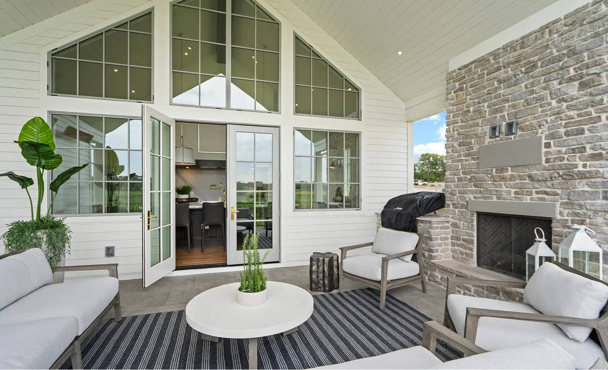 Covered outdoor patio with cushioned chairs, a round table, stone fireplace, and open glass doors to the kitchen.