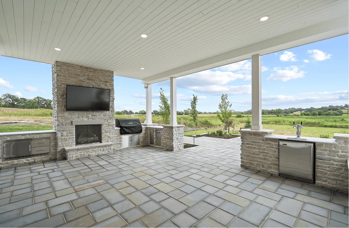Covered outdoor patio with stone fireplace, TV, built-in grill, sink, and open views of greenery.