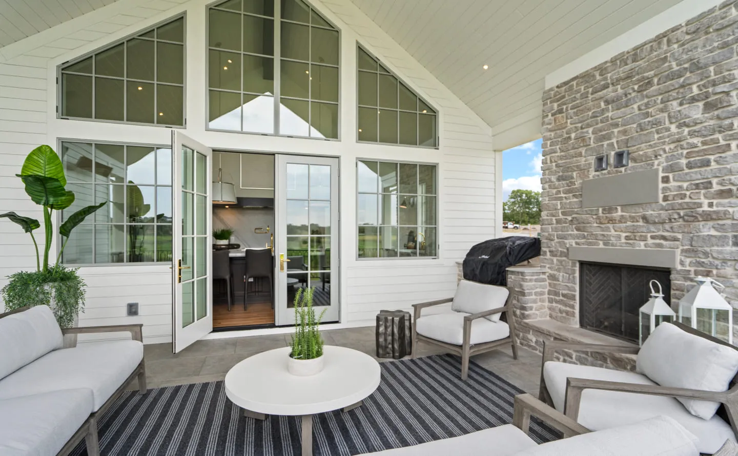 Covered patio with gray cushioned seats, white round table, black striped rug, and stone fireplace.