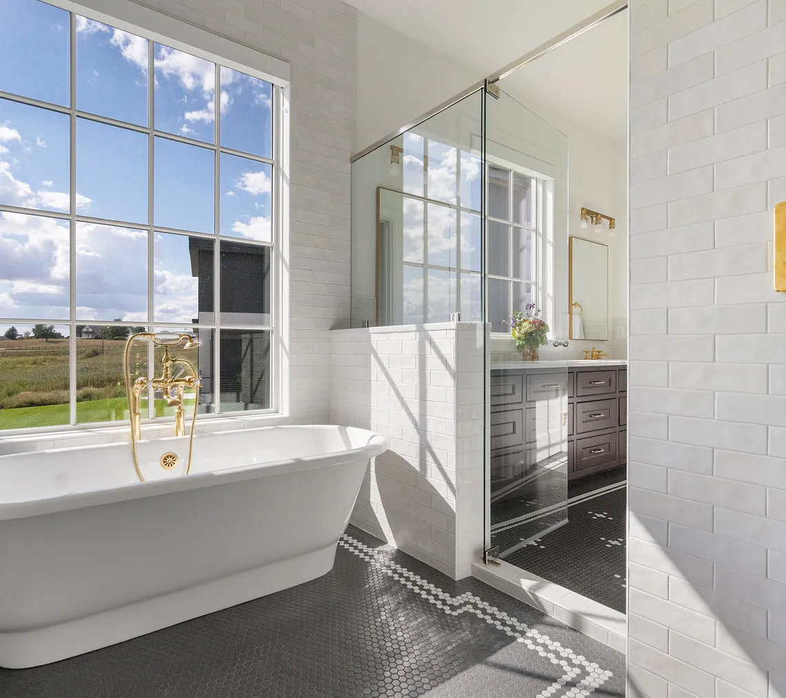 Bright bathroom with a white freestanding tub, large window, glass shower, and dark hex tile floor.