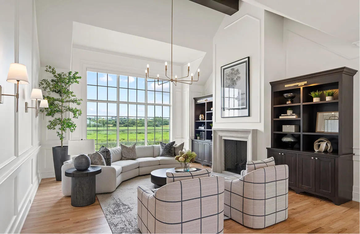 Bright living room with large window overlooking green landscape, gray curved sofa, patterned chairs, and fireplace.