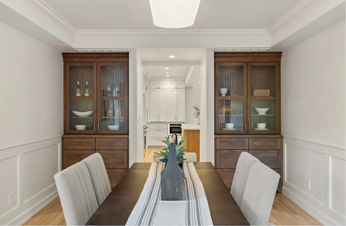 Dining room with dark wood table, six beige chairs, two wooden cabinets, and view into white kitchen.
