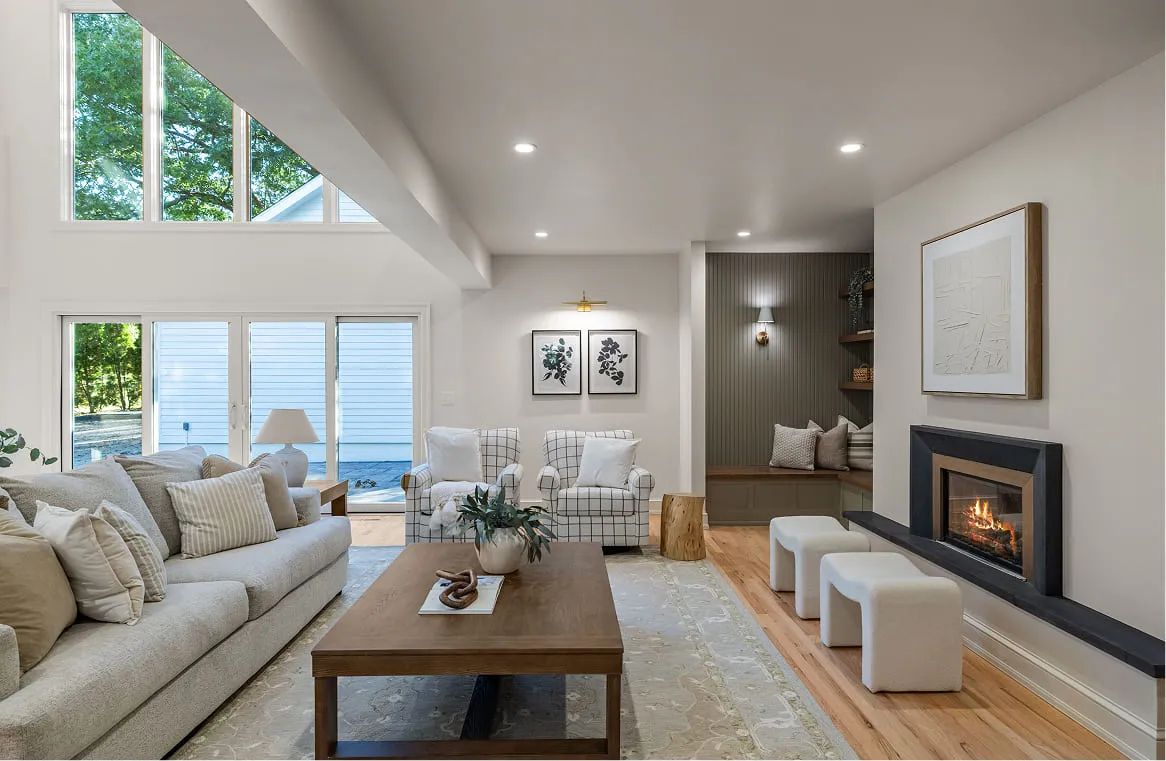 Bright modern living room with fireplace, two checkered armchairs, beige sofa, wooden table, and large windows.