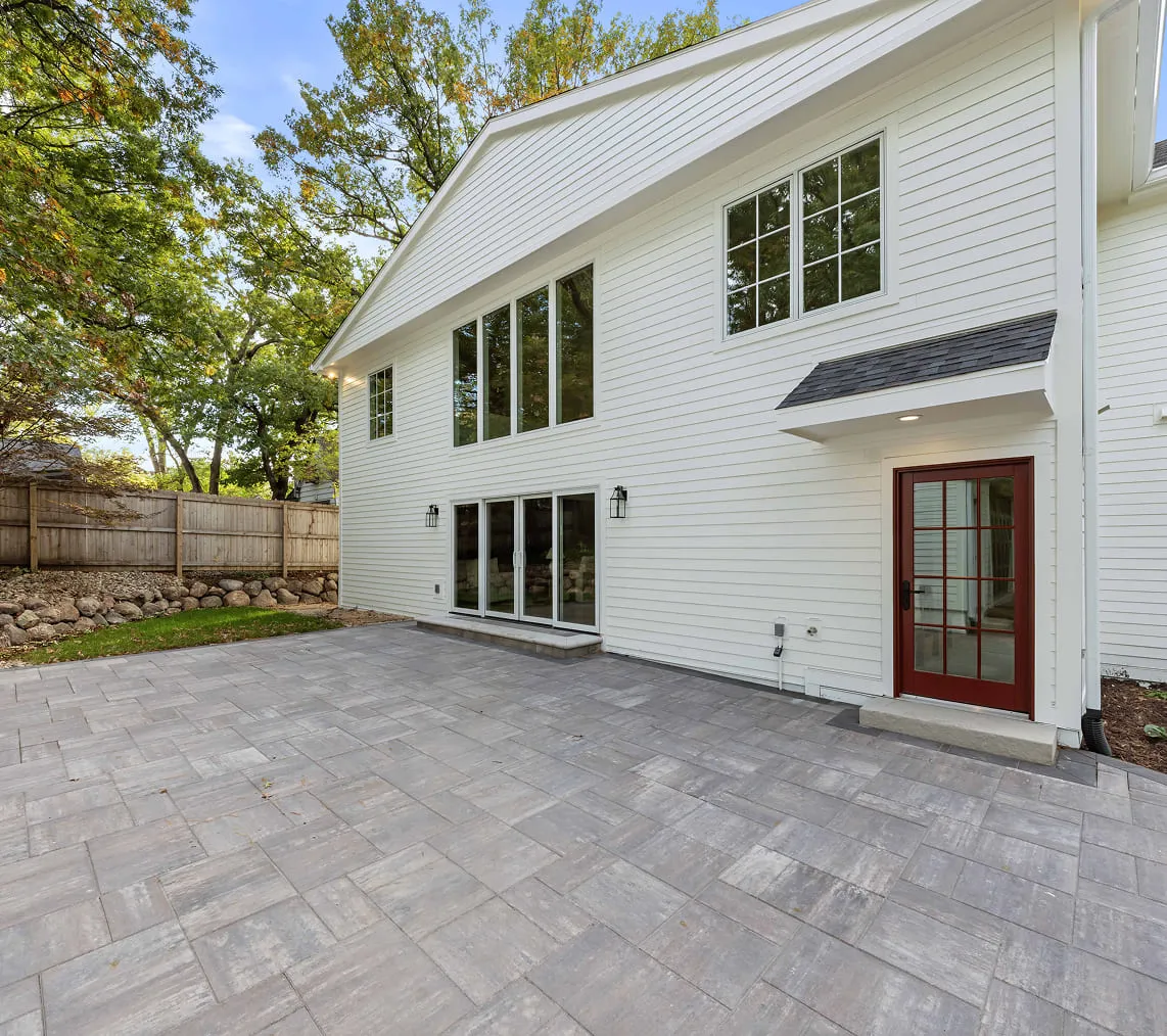 Backyard patio with gray stone tiles outside a white house with large windows and a red door.