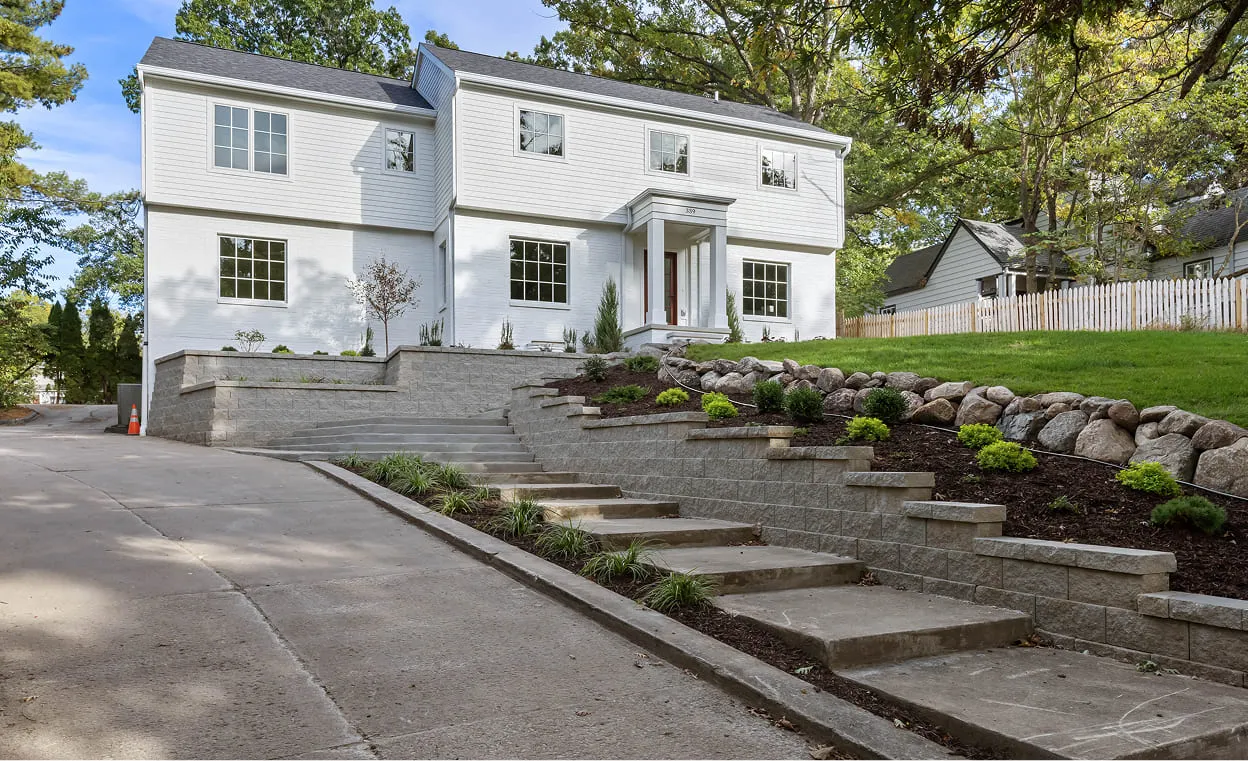 White two-story house with a concrete driveway, tiered stone retaining walls, and landscaped plants.