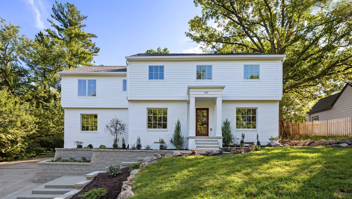 White two-story house with multiple windows, front door, stone retaining walls, and green lawn.