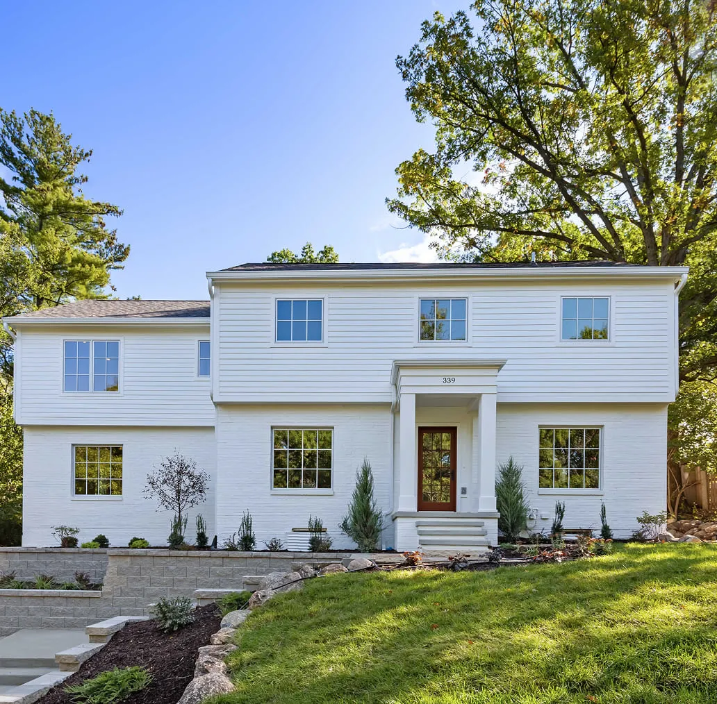 White two-story house with large windows, front steps, and landscaping including trees and grass.