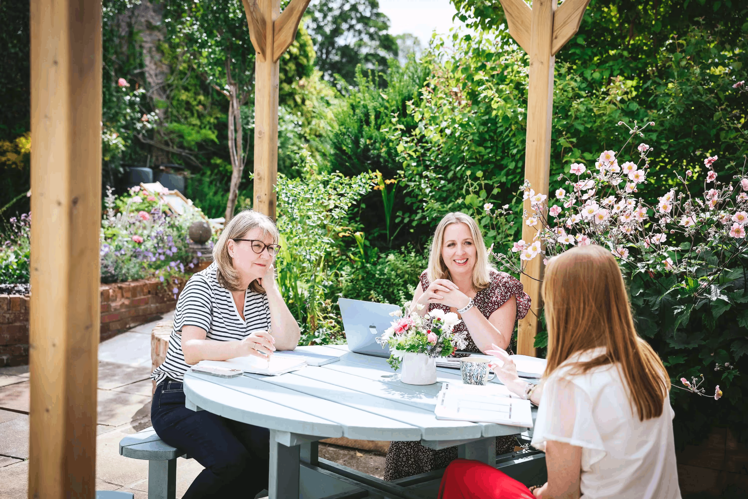Three women chatting around a garden table under a wooden pergola