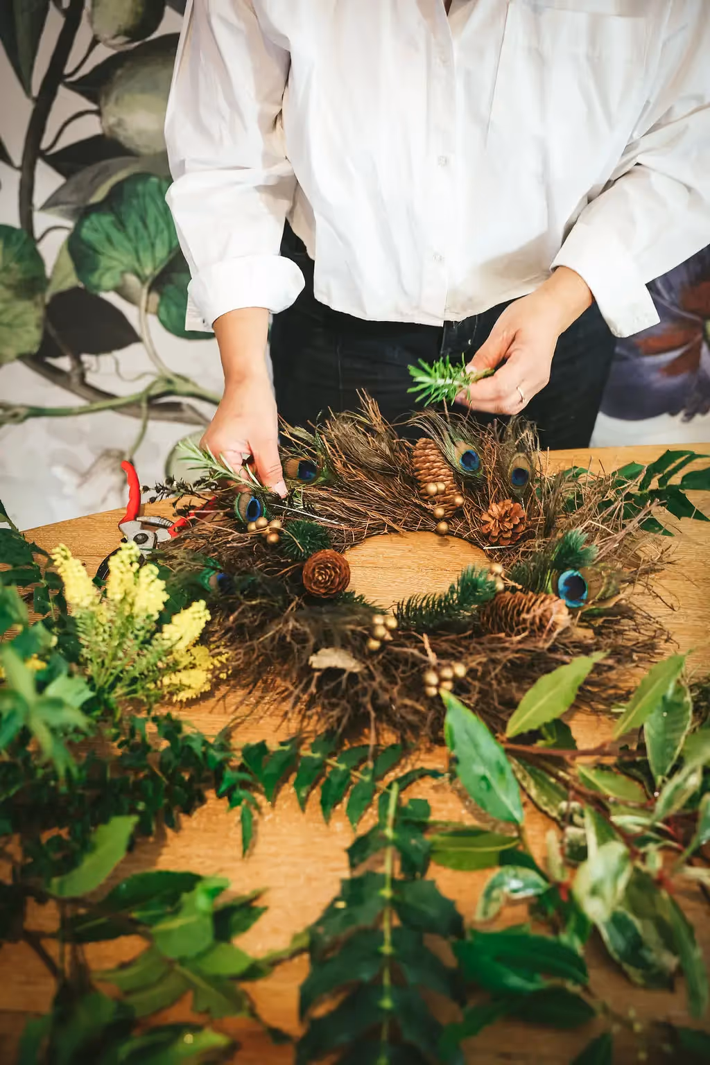 Hands arranging a natural foliage wreath with pinecones and peacock feathers