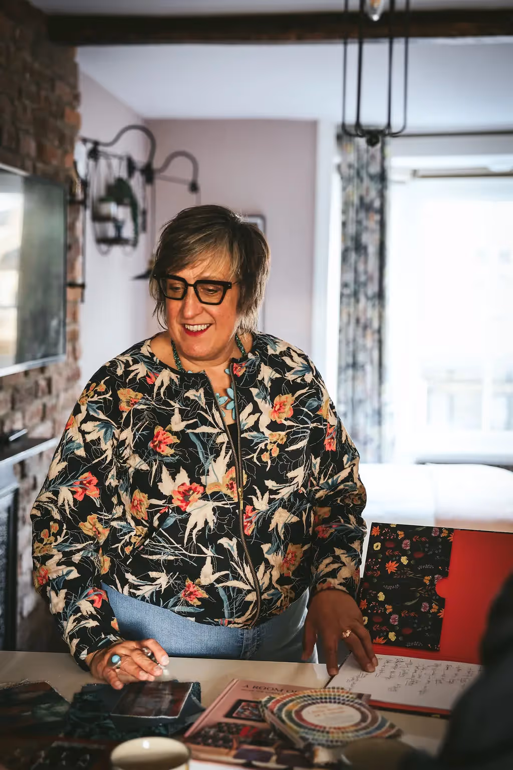 Smiling woman in a floral jacket browsing fabric samples by a window