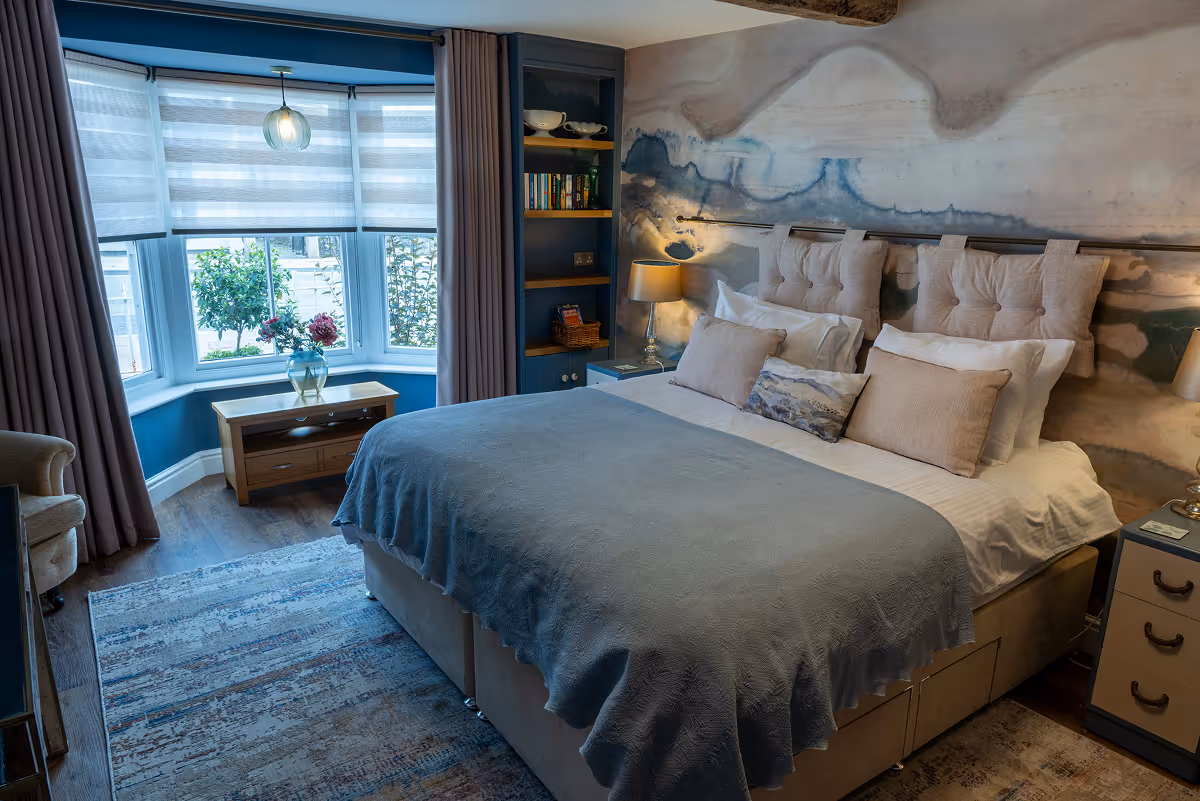 Snug bedroom with grey throw, cushions and a sunlit bay window