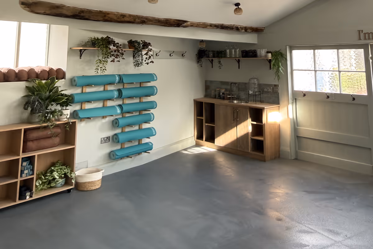 Bright yoga room with teal mats on wall rack, wooden shelves, plants, and a small kitchenette.