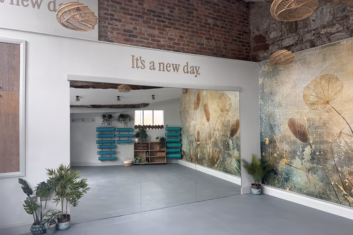 Empty studio room with large wall mirror, floral mural, plants, and ‘It’s a new day’ sign on wall.