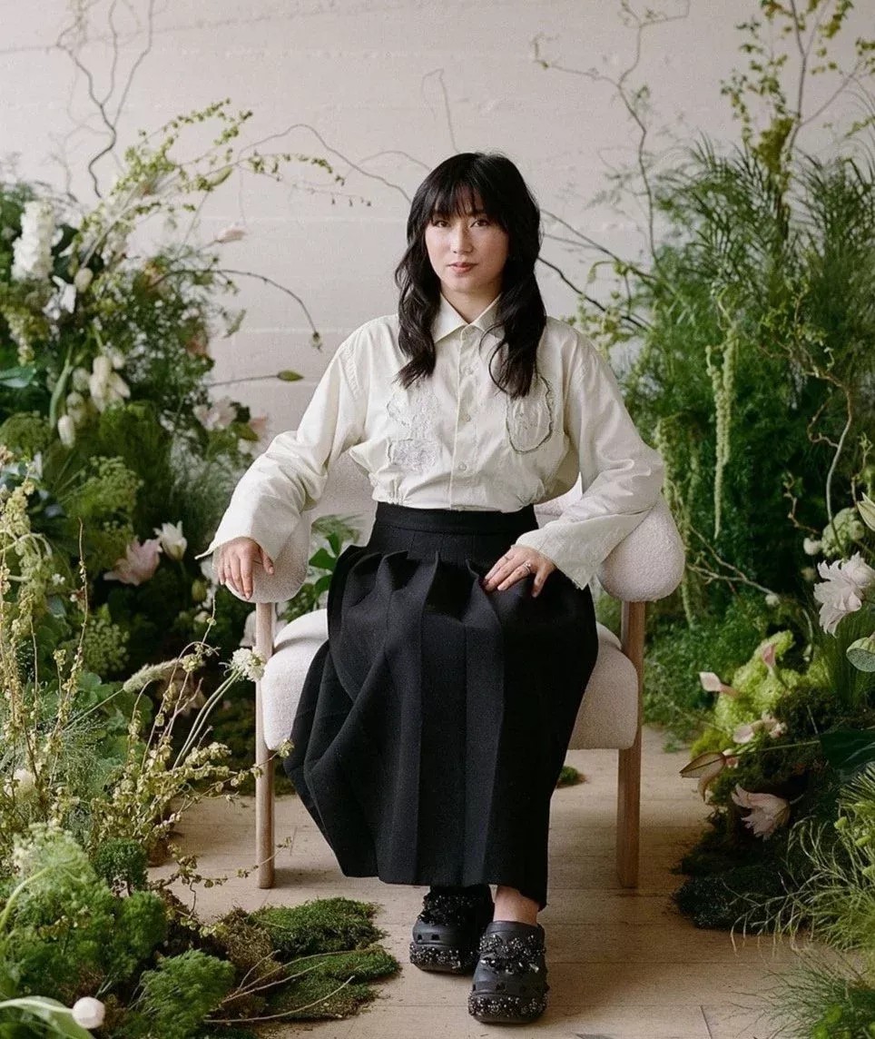 Atelier Amarise client portrait — woman in white shirt and black pleated skirt seated among lush greenery and white florals